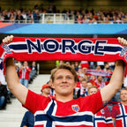 Fanshoppen Norge red, white, and blue horizontal scarf with NORWAY text and Norwegian flag details. Young man wearing a Norway jersey holds it up.